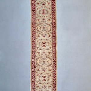 May include: A long, narrow rug with a cream background and a red border. The rug has a floral pattern in the center and a repeating pattern along the border.