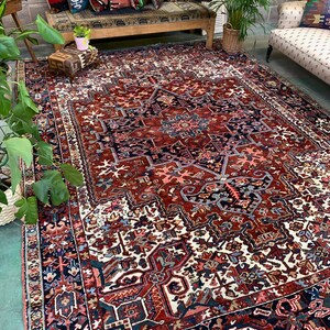 May include: A large, intricately patterned rug with a red, blue, and white color scheme. The rug has a central medallion and a border with geometric designs. The rug is laid out on a concrete floor in a room with a wooden bench and plants.
