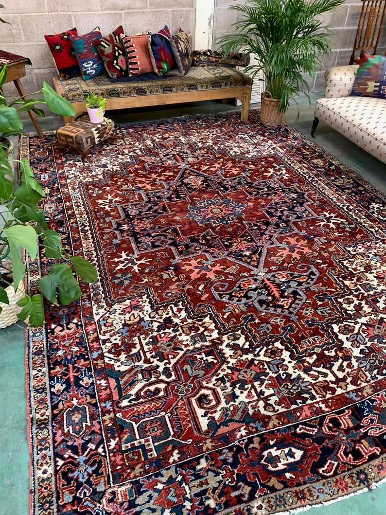 May include: A large, intricately patterned rug with a red, blue, and white color scheme. The rug has a central medallion and a border with geometric designs. The rug is laid out on a concrete floor in a room with a wooden bench and plants.