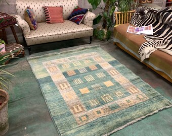 Op de afbeelding: Een groen en beige tapijt met een geometrisch ontwerp. Het tapijt ligt op een betonnen vloer voor een bank en een sofa.