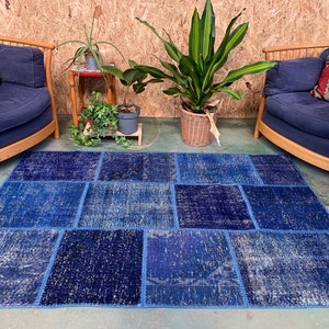 Op de afbeelding: Een blauw patchwork tapijt met een verweerde look, geplaatst op een houten vloer voor twee fauteuils met kussens. Het tapijt is gemaakt van verschillende tinten blauw denim stof en heeft een unieke, handgemaakte uitstraling.