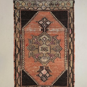 Op de afbeelding: Een klein, rechthoekig tapijt met een traditioneel Turks ontwerp. Het tapijt heeft een donkerbruine rand met een centraal medaillon in tinten rood, groen en zwart. Het tapijt heeft een franjerand.