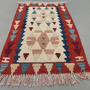 Puede incluir: Una alfombra kilim turca tradicional con un patrón geométrico en tonos rojos, azules y crema. La alfombra tiene un borde de flecos.