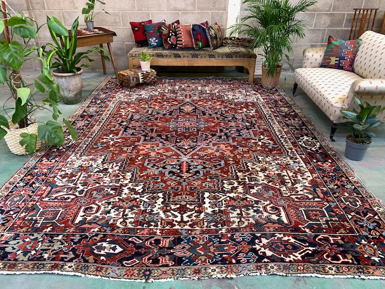 May include: A large, intricately patterned rug with a red, white, and blue color scheme. The rug has a central medallion and a border with geometric designs. The rug is laid out on a concrete floor in a room with plants and furniture.