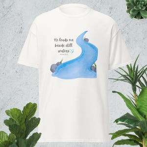 Może przedstawiać: Biała koszulka z akwarelowym wzorem niebieskiej rzeki, szarych kamieni i zielonych akcentów. Tekst "He leads me beside still waters" jest wydrukowany nad rzeką, ze znakiem serca. Poniżej wydrukowano Psalm 23:2.