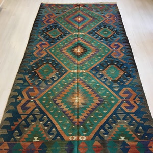 May include: A rectangular, hand-woven rug with a geometric pattern. The rug features a central diamond design in teal, orange, and purple, framed by a dark blue border. The rug's design includes repeating diamond and triangle shapes.