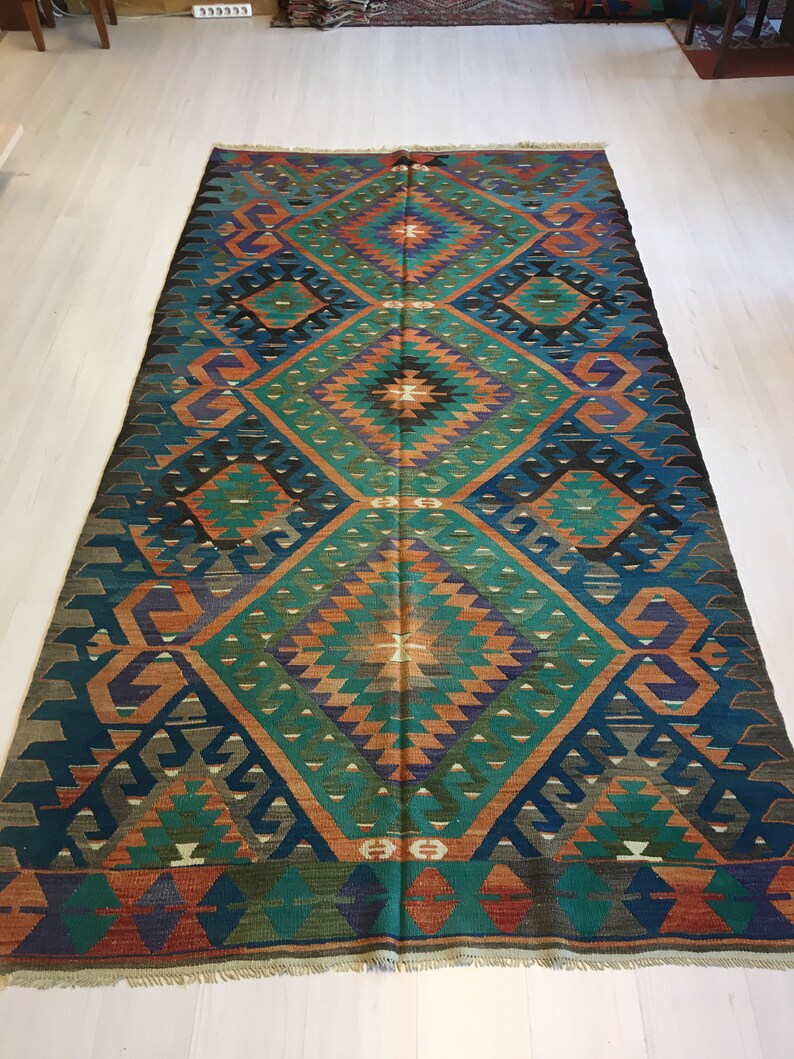 May include: A rectangular, hand-woven rug with a geometric pattern. The rug features a central diamond design in teal, orange, and purple, framed by a dark blue border. The rug's design includes repeating diamond and triangle shapes.