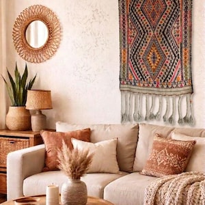 Puede incluir: Un salón con un sofá beige, cojines y una mesa de centro de madera. Un tapiz de pared tejido con un diseño geométrico en tonos rojos, negros y naranjas cuelga sobre el sofá. Un espejo redondo y plantas completan la decoración.