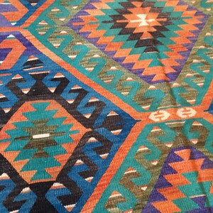 May include: Close-up of a colorful woven rug with a geometric pattern. The rug features a diamond design with teal, orange, blue, and black shapes. The rug's texture is visible, suggesting a handmade or artisanal quality.