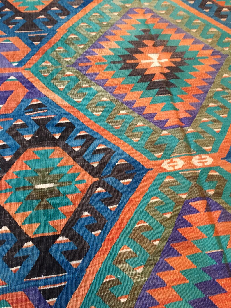 May include: Close-up of a colorful woven rug with a geometric pattern. The rug features a diamond design with teal, orange, blue, and black shapes. The rug's texture is visible, suggesting a handmade or artisanal quality.