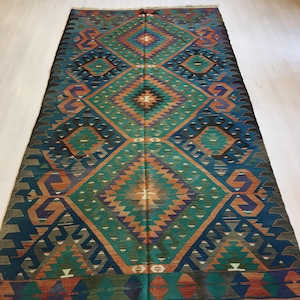 May include: A rectangular, hand-woven rug with a geometric pattern. The rug features a central diamond design in shades of teal, orange, and black, with a border of teal and brown. The rug is placed on a light-colored wooden floor.