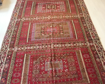 May include: A rectangular, red and brown patterned rug with a central design of repeating squares. The rug features intricate geometric patterns and a fringed edge. The color palette includes shades of red, brown, green, and beige.