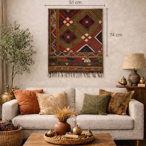 Puede incluir: Un tapiz de pared rectangular tejido, de 51 cm de ancho y 74 cm de largo, con un patrón geométrico en rojo, verde y azul. La alfombra tiene flecos en la parte inferior y cuelga sobre un sofá beige con cojines decorativos. La habitación tiene un ambiente cálido y acogedor.