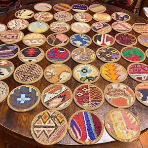 May include: A large collection of circular coasters featuring a variety of woven textile patterns in diverse colors including red, blue, yellow, green, and brown. Each coaster has a distinct geometric or floral design with a finished edge.