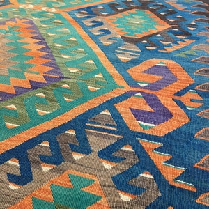 May include: A colorful, geometric patterned rug with a mix of teal, orange, blue, purple, and brown hues. The rug features a repeating design of angular shapes and patterns, creating a vibrant and textured surface. The rug is likely made of woven fabric.