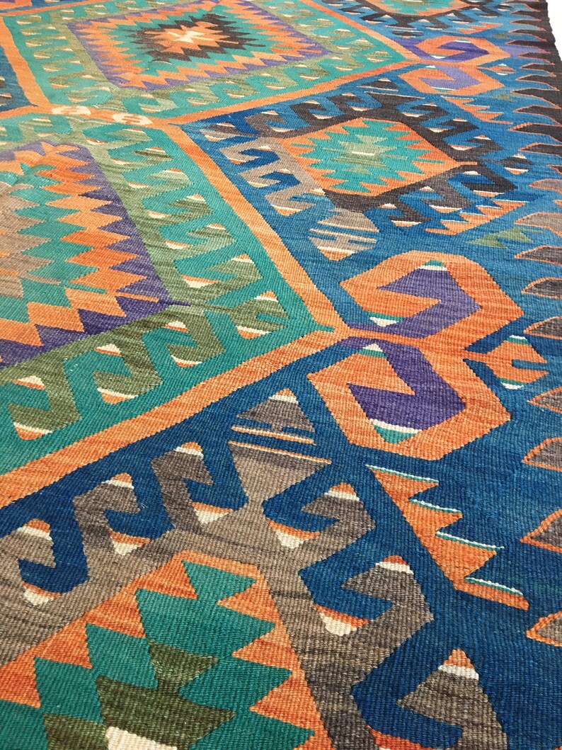 May include: A colorful, geometric patterned rug with a mix of teal, orange, blue, purple, and brown hues. The rug features a repeating design of angular shapes and patterns, creating a vibrant and textured surface. The rug is likely made of woven fabric.