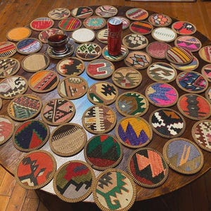 Può includere: Una collezione di sottobicchieri rotondi e colorati con intricati motivi tessuti, disposti su un tavolo di legno. I sottobicchieri presentano vari disegni in tonalità di rosso, blu, verde e giallo. Una lattina di soda e un bicchiere di tè sono anche sul tavolo.