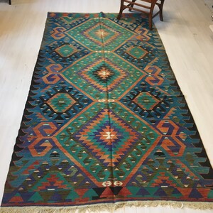 May include: A rectangular, hand-woven rug with a geometric pattern. The rug features a central diamond design in teal, orange, and purple hues, framed by a dark blue border. The rug has a fringed edge and is placed on a light-colored wooden floor.