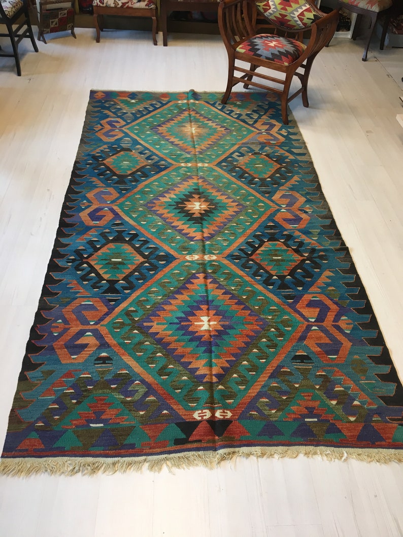 May include: A rectangular, hand-woven rug with a geometric pattern. The rug features a central diamond design in teal, orange, and purple hues, framed by a dark blue border. The rug has a fringed edge and is placed on a light-colored wooden floor.