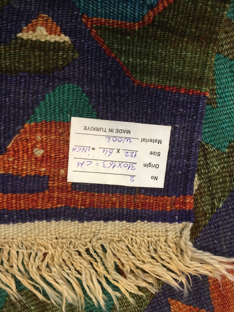 May include: A hand-woven wool rug with a geometric pattern in shades of blue, green, orange, and cream. The rug measures 122 x 64 inches and has a fringed edge. The tag indicates it was made in Turkiye.