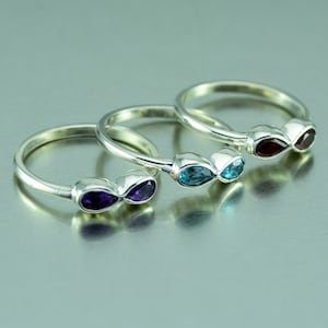 Könnte beinhalten: Drei Silberringe mit Unendlichkeits-Symbolen. Jeder Ring ist mit einem anderen Edelstein besetzt: Amethyst, blauer Topas und Granat.