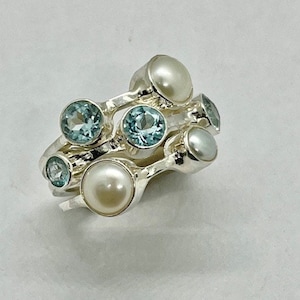 May include: A silver ring with multiple bands featuring blue gemstones and white pearls. The ring is designed with a unique, organic shape.