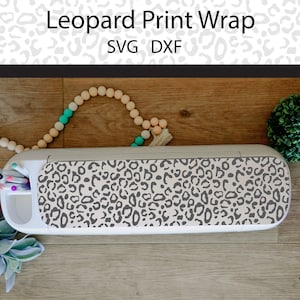 Puede incluir: Una máquina de manualidades blanca con un diseño de estampado de leopardo en la parte superior. La máquina está sobre una superficie de madera con una cadena de cuentas y una pequeña planta en maceta.