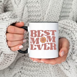 May include: White ceramic mug with a black rim and handle. The mug has a pink and yellow graphic that says "Best Mom Ever".