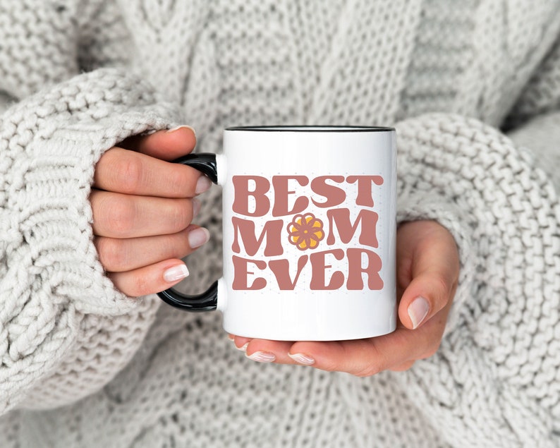 May include: White ceramic mug with a black rim and handle. The mug has a pink and yellow graphic that says "Best Mom Ever".