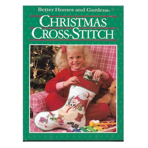 May include: A book titled "Better Homes and Gardens Christmas Cross-Stitch" shows a young girl holding a Christmas stocking filled with cross-stitch designs of toys and a snowman.