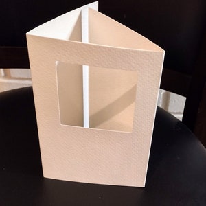 May include: A cream-colored, triangular paper box with a rectangular window cut out of the front. The box has a textured surface and is open at the top. The box is designed to hold a small item.