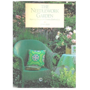 Op de afbeelding: Een boekomslag getiteld "The Needlework Garden" met een groene rieten stoel en een kussen met een patroon. De stoel staat in een tuin met bloemen en groen. Het boek is van Jane Iles.