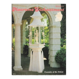 Puede incluir: Una fuente de macramé blanca con un diseño en capas, con una estatua central y adornos colgantes. La portada del libro dice "Macramé Enchantment" en escritura rosa, con "Book 4" arriba. El título "Fountain of the Orient" está en la parte inferior.