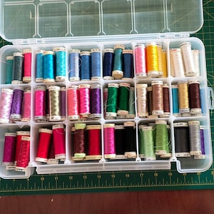 May include: A clear plastic storage container filled with an assortment of colourful sewing thread spools. The spools are arranged in rows and columns, featuring a wide spectrum of colours including pink, blue, green, yellow, brown, and black.