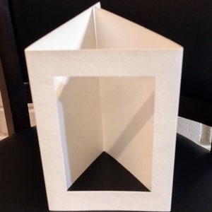 May include: A white, geometric paper box with a rectangular opening and a triangular top. The box is constructed from thick, textured paper and has a clean, minimalist design. The interior is black, creating a contrast with the white exterior.