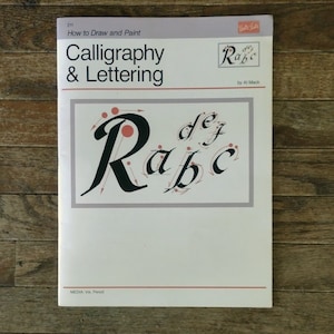 May include: Book cover for "Calligraphy & Lettering" by Al Mack. The cover features a pink and black illustration of the letters "Rabe" with arrows showing the direction of strokes.