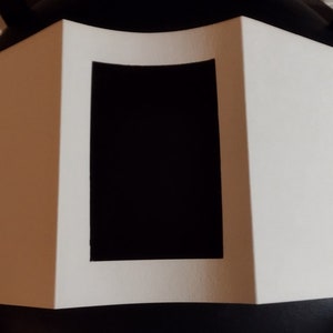 May include: A white, folded paper display with a black rectangular cutout in the center. The display is sitting on a black surface, with a wooden floor visible in the background.