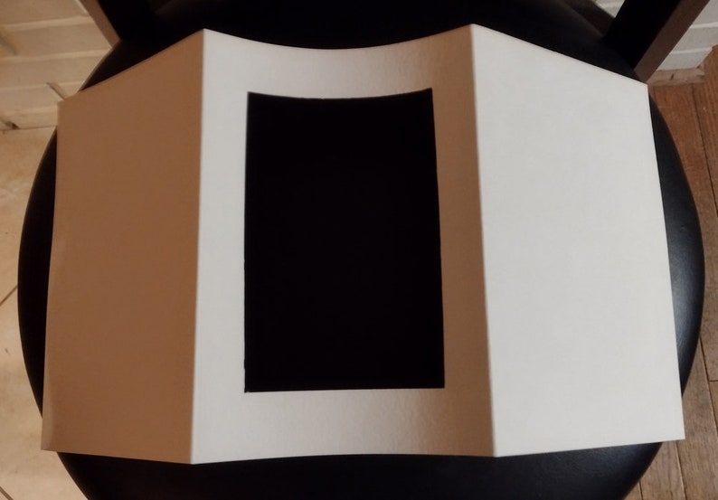 May include: A white, folded paper display with a black rectangular cutout in the center. The display is sitting on a black surface, with a wooden floor visible in the background.