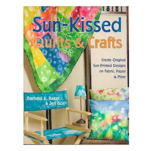 以下が含まれることがあります： バーバラ・K・ベーカーとジェリ・ボーによる「Sun-Kissed Quilts & Crafts」というタイトルの本。表紙には、カラフルなパッチワークキルト、ティール色の布製シートが付いたディレクターズチェア、ティール色のシェードが付いた小さなランプ、2つの装飾用クッションが写っています。