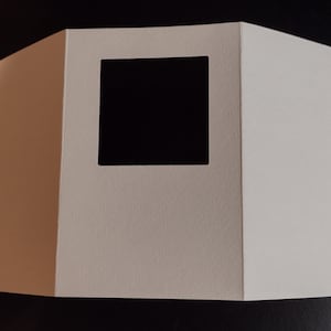 May include: A white, folded paper display with a square cutout. The display is made of thick paper and has a geometric shape. The cutout reveals a black interior, creating a contrast.