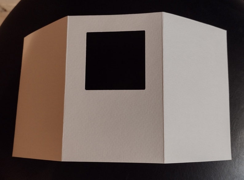 May include: A white, folded paper display with a square cutout. The display is made of thick paper and has a geometric shape. The cutout reveals a black interior, creating a contrast.
