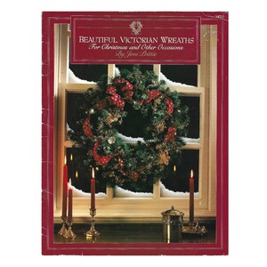 Peut inclure: Un livre intitulé Beautiful Victorian Wreaths For Christmas and Other Occasions par Joni Prittie. La couverture présente une bordure rouge foncé et une image centrale d'une couronne de pin verte avec des rubans rouges suspendue à une fenêtre enneigée, avec des bougies rouges.