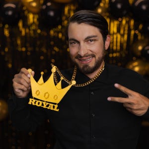 May include: A gold crown pendant necklace with the word "ROYAL" on it. The necklace is worn by a person with a beard and a black shirt.