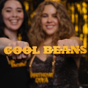 May include: Two women are holding up a gold-colored sign that says "COOL BEANS". The women are wearing sparkly outfits and are smiling.