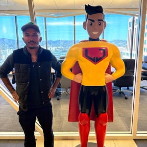 May include: A man in a green and black baseball cap stands next to a large, cartoon superhero figure. The superhero is wearing a yellow suit with a red cape and black boots. The superhero's chest has a red and black logo with the letters "LJ".