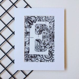 May include: A black and white drawing of the letter "E" surrounded by intricate floral patterns. The letter is outlined in white and the background is filled with detailed designs.