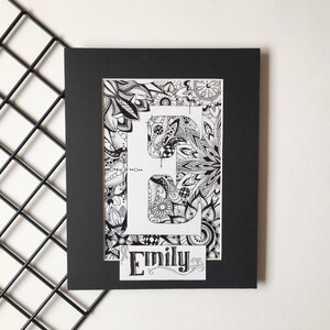 May include: Black and white framed print of the letter "E" with a decorative floral pattern. The name "Emily" is written in a decorative font below the letter.
