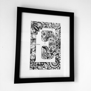 May include: Black and white framed print of the letter "E" surrounded by intricate floral designs.