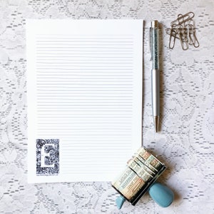 May include: A white sheet of lined paper with a black and white illustration of the letter "E" in the lower left corner. A silver pen with glitter inside is on the right side of the paper. A blue and white rubber stamp with numbers and letters is in the lower right corner.
