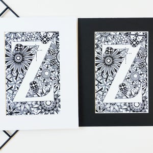 May include: Two framed prints of a black and white illustration of the letter Z surrounded by intricate patterns. The print on the left is framed in white, and the print on the right is framed in black.